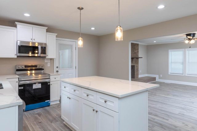 Kitchen and living room remodel featuring large island and hanging lights with white countertops, gray backsplash, and light wood flooring